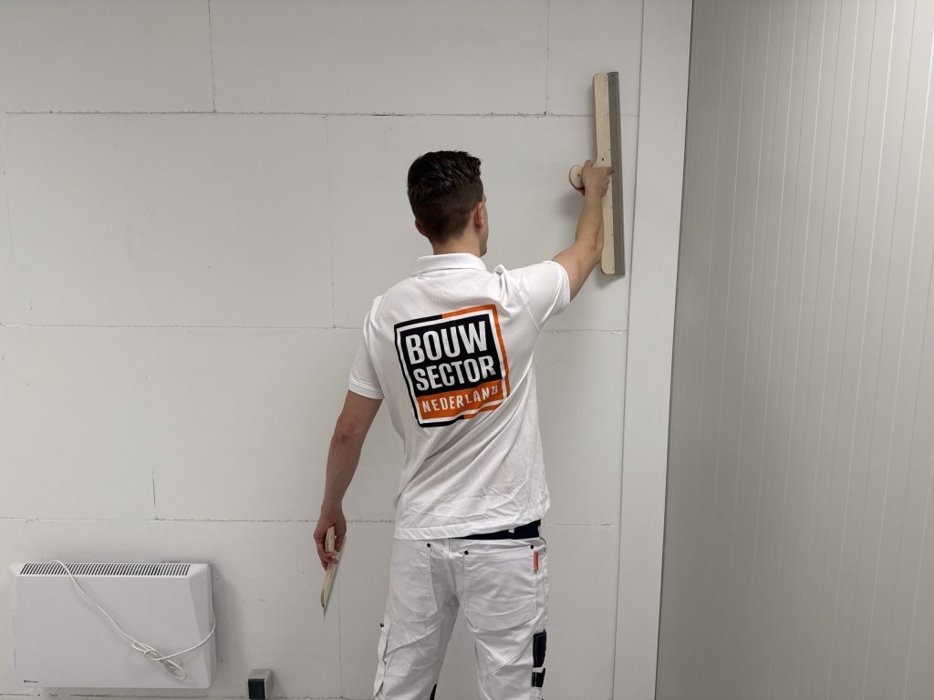 Stukadoor van Bouwsector Nederland zet bovenaan een wand vlak met een lange spaan, een pleisterspaan in de andere hand