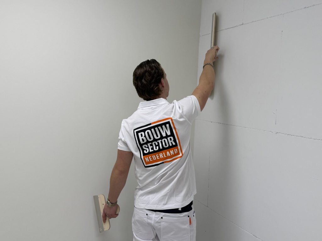 Stukadoor van Bouwsector Nederland strijkt met een lange spaan een wand in de hoek van een kamer glad